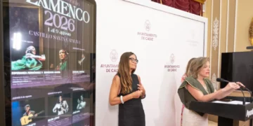 Two women at a formal event: one at a podium speaking, the other standing beside a white backdrop with Ayuntamiento de Cádiz logos.