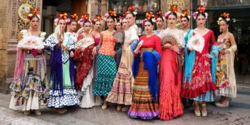 Group of women in vibrant flamenco dresses with flowers in hair, posing on a decorative street.