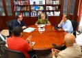 Six people sit around a round wooden conference table in a formal office, discussing documents and taking notes.