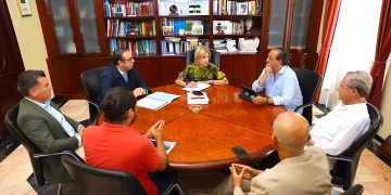 Six people sit around a round wooden conference table in a formal office, discussing documents and taking notes.