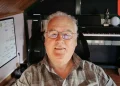 Man in plaid shirt and glasses smiles at the camera in a home studio; a piano, plant, and large monitor are visible behind him; EXPO MÚSICA badge top-right.