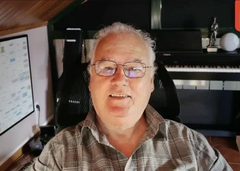 Man in plaid shirt and glasses smiles at the camera in a home studio; a piano, plant, and large monitor are visible behind him; EXPO MÚSICA badge top-right.